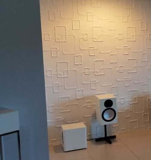 White bookshelf speaker on a black stand beside a small white subwoofer against a textured feature wall