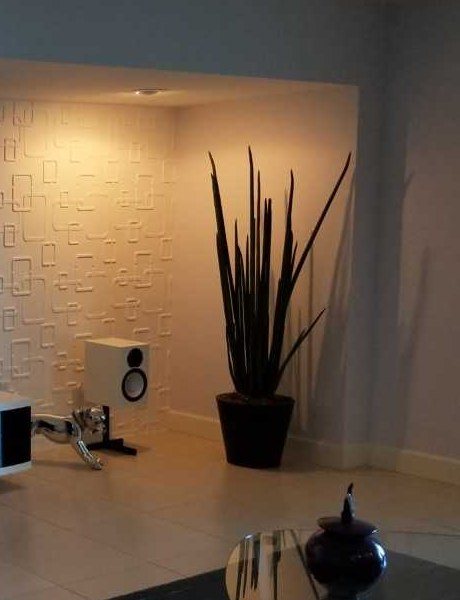 White bookshelf speaker on a black stand beside a tall plant against a lit textured feature wall