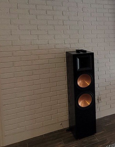 A black floorstanding tower speaker with copper woofers standing out from a painted white brick wall